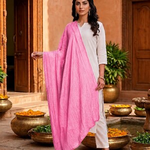May include: A woman wearing a white outfit with a bright pink scarf. The scarf is draped over the shoulders and arms. The outfit is paired with black heels. The background features a courtyard with golden pots and plants.