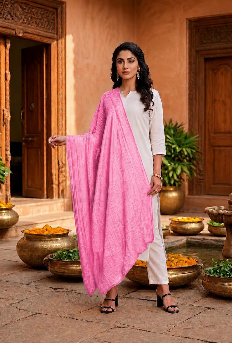 May include: A woman wearing a white outfit with a bright pink scarf. The scarf is draped over the shoulders and arms. The outfit is paired with black heels. The background features a courtyard with golden pots and plants.