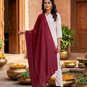 May include: A woman wearing a white outfit with a long, flowing burgundy scarf. The outfit includes a white top and pants. The background features a courtyard with decorative pots and plants.