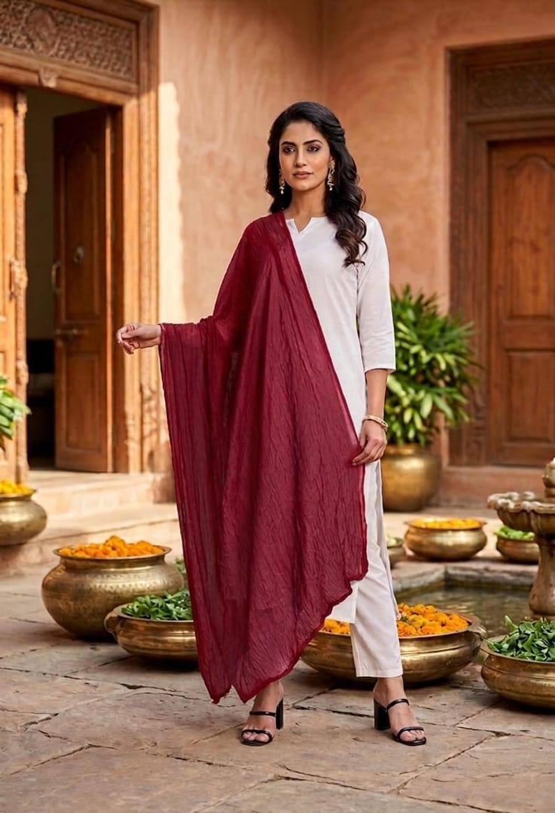 May include: A woman wearing a white outfit with a long, flowing burgundy scarf. The outfit includes a white top and pants. The background features a courtyard with decorative pots and plants.