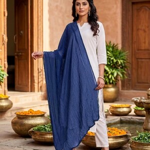 May include: A woman wearing a white outfit with a long, flowing, navy blue scarf. The outfit includes a white top and pants. The woman is wearing black heels. The background features a courtyard with decorative pots and wooden doors.