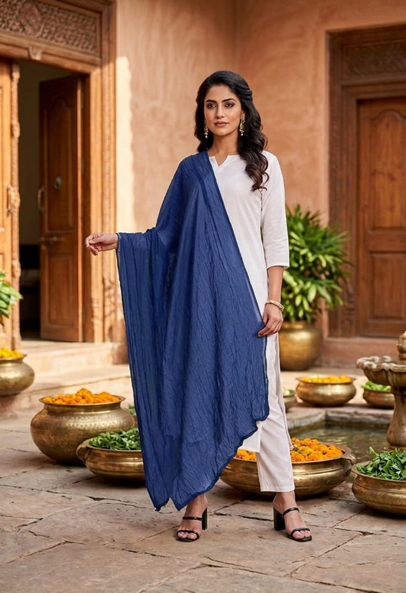 May include: A woman wearing a white outfit with a long, flowing, navy blue scarf. The outfit includes a white top and pants. The woman is wearing black heels. The background features a courtyard with decorative pots and wooden doors.