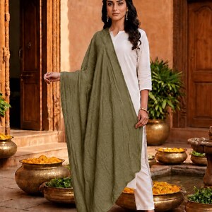 May include: A woman wearing a white outfit with a long olive green scarf. The outfit includes a long-sleeved top and pants. The background features ornate wooden doors, stone flooring, and decorative pots.