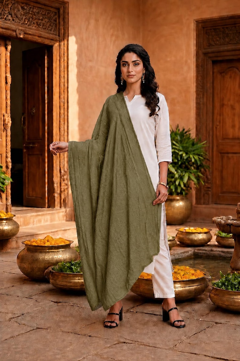May include: A woman wearing a white outfit with a long olive green scarf. The outfit includes a long-sleeved top and pants. The background features ornate wooden doors, stone flooring, and decorative pots.