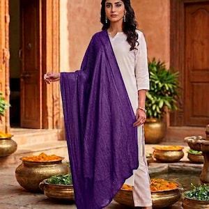 May include: A woman wearing a white outfit with a long, flowing, deep purple scarf. The scarf is draped over the shoulders and arms. The background includes gold pots with orange flowers and green plants.
