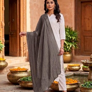 May include: A woman wearing a white outfit with a gray scarf. The scarf is draped over the shoulders and has a textured appearance. The woman is standing in front of a stone patio with decorative pots.