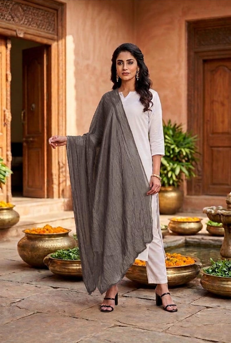 May include: A woman wearing a white outfit with a gray scarf. The scarf is draped over the shoulders and has a textured appearance. The woman is standing in front of a stone patio with decorative pots.