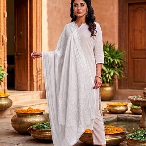 May include: A woman wearing a white kurta and matching pants, paired with a white scarf. The outfit is set against a backdrop of a courtyard with bronze pots filled with flowers and greenery.
