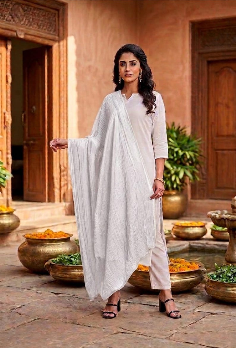 May include: A woman wearing a white kurta and matching pants, paired with a white scarf. The outfit is set against a backdrop of a courtyard with bronze pots filled with flowers and greenery.