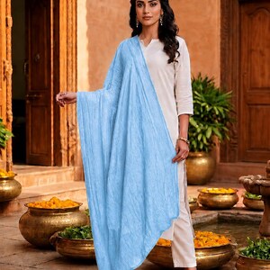May include: A woman wearing a light blue scarf draped over a cream-colored outfit. The scarf has a textured appearance. The outfit includes a top and pants. The background features a courtyard with decorative pots and wooden doors.