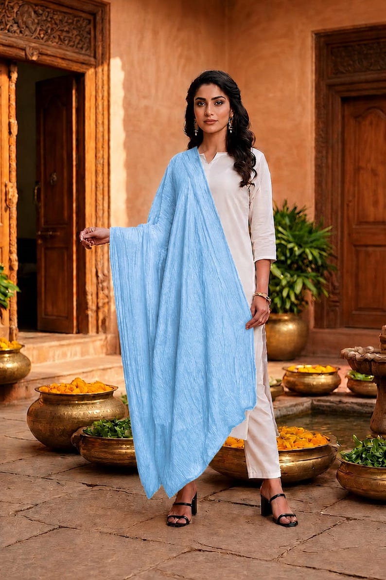 May include: A woman wearing a light blue scarf draped over a cream-colored outfit. The scarf has a textured appearance. The outfit includes a top and pants. The background features a courtyard with decorative pots and wooden doors.