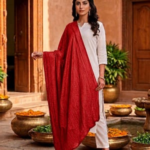 May include: A woman wearing a white outfit with a vibrant red shawl. The shawl is draped over the shoulders and has a textured appearance. The outfit is paired with black heels. The background features a courtyard with decorative pots.