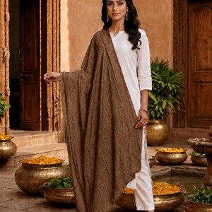 May include: A woman wearing a white outfit with a brown shawl. The shawl is draped over the shoulders and arms. The background includes wooden doors, pots with yellow flowers, and a water feature.