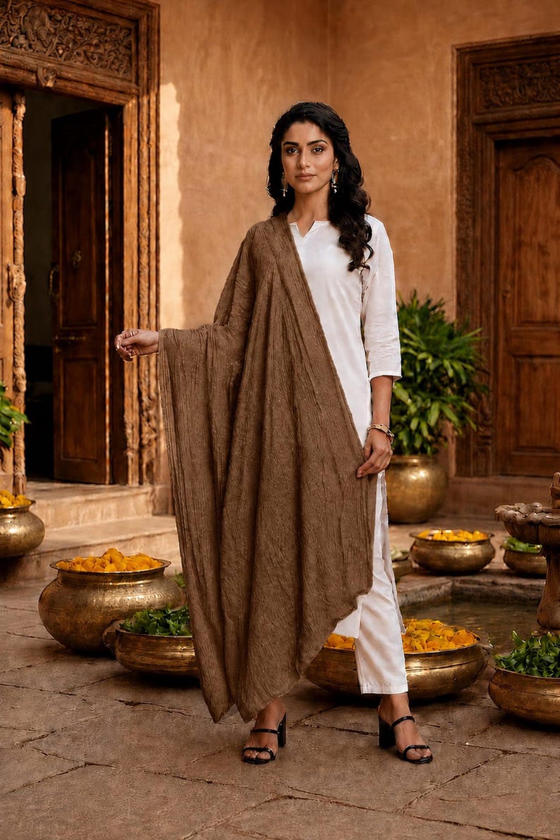 May include: A woman wearing a white outfit with a brown shawl. The shawl is draped over the shoulders and arms. The background includes wooden doors, pots with yellow flowers, and a water feature.