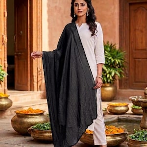 May include: A woman wearing a white outfit with a long black scarf. The scarf is draped over the shoulders and arms. The background features a courtyard with decorative pots and plants.
