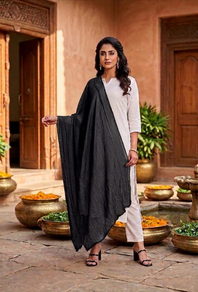 May include: A woman wearing a white outfit with a long black scarf. The scarf is draped over the shoulders and arms. The background features a courtyard with decorative pots and plants.