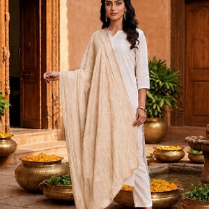 May include: A woman wearing a white kurta and pants with a beige scarf. She is standing in front of a traditional Indian building. The scene includes gold pots with yellow flowers and green plants.