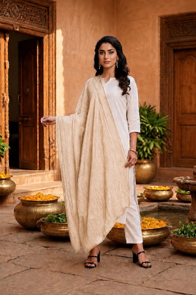 May include: A woman wearing a white kurta and pants with a beige scarf. She is standing in front of a traditional Indian building. The scene includes gold pots with yellow flowers and green plants.