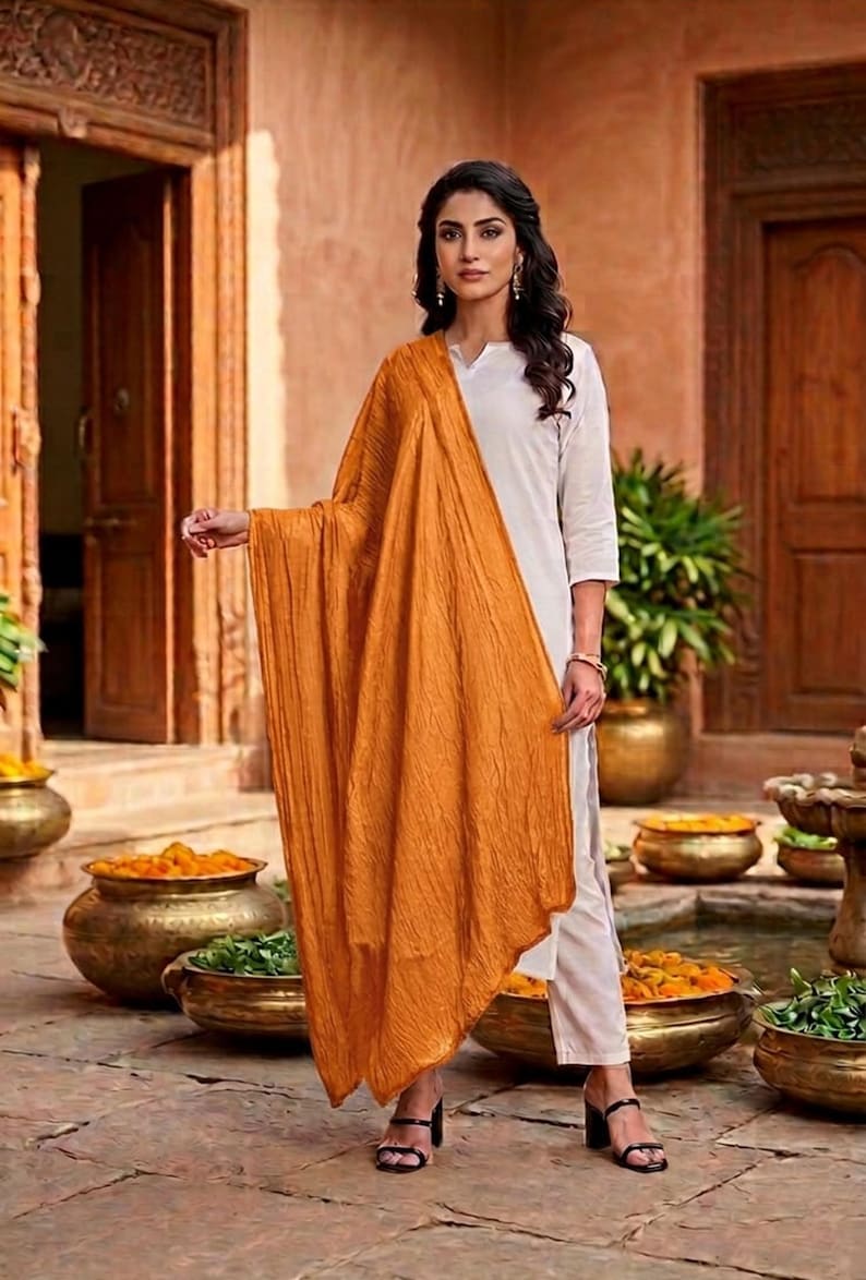 May include: A woman wearing a white outfit with an orange shawl. The shawl is draped over the shoulders and arms. The background features a courtyard with decorative pots and plants.
