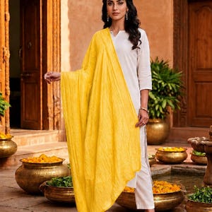 May include: A woman wearing a white outfit with a bright yellow scarf. The scarf is draped over the shoulders and arms. The outfit includes white pants and a white top. The woman is wearing black heels.