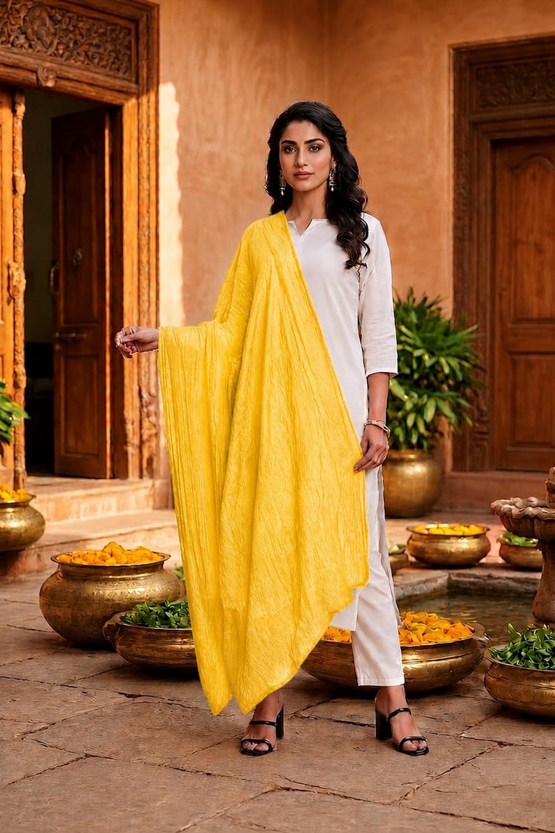 May include: A woman wearing a white outfit with a bright yellow scarf. The scarf is draped over the shoulders and arms. The outfit includes white pants and a white top. The woman is wearing black heels.