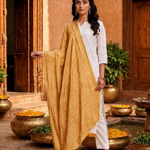 May include: A woman wearing a white outfit with a mustard-colored shawl. The outfit includes a long-sleeved top and pants. The shawl is draped over the shoulders. The background features a courtyard with bronze pots and wooden doors.