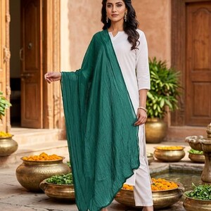May include: A woman wearing a white tunic and pants, paired with a flowing, emerald green scarf. The outfit is accessorized with black sandals. The background features a courtyard with decorative pots and a water feature.