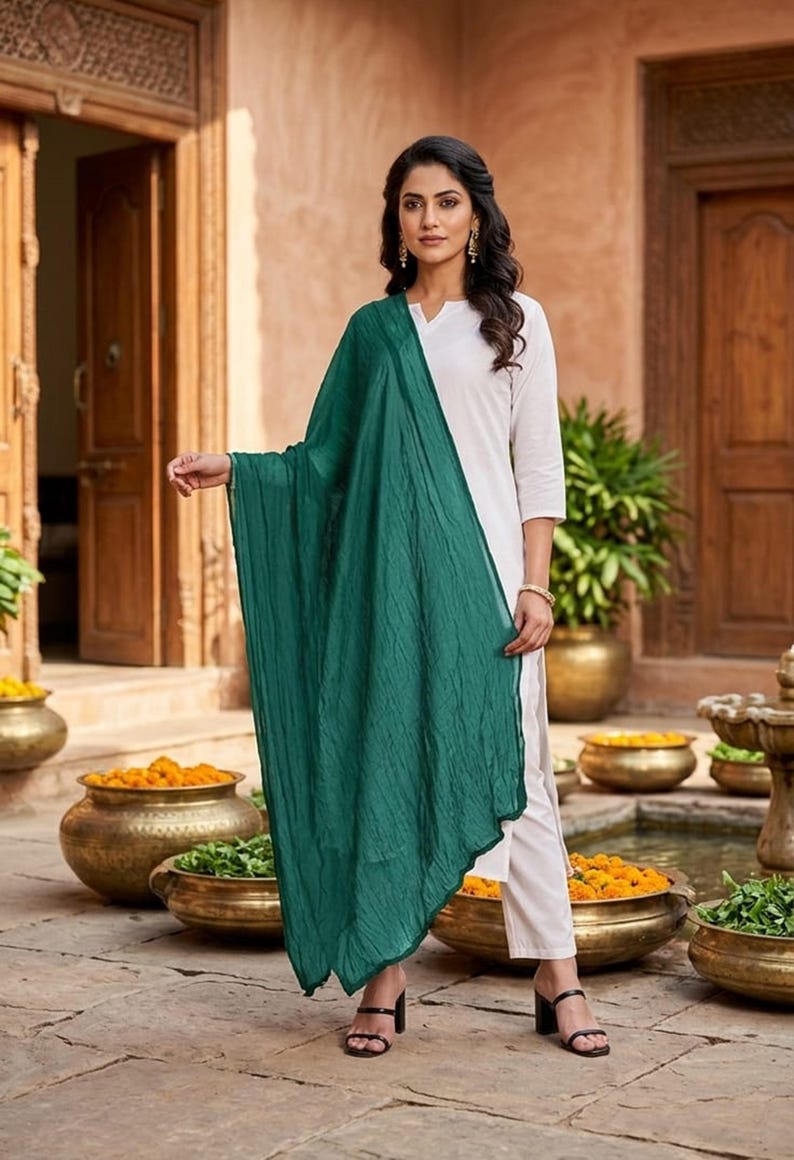 May include: A woman wearing a white tunic and pants, paired with a flowing, emerald green scarf. The outfit is accessorized with black sandals. The background features a courtyard with decorative pots and a water feature.