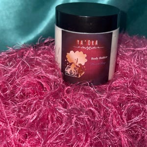 May include: A white jar of body butter with a black lid, labelled "YA'ORA Body Butter". The jar sits on a bed of bright pink, fluffy material against a teal background. The label has a maroon background with a graphic.