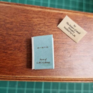 Puede incluir: Un pequeño libro azul con el texto "Part of L.M.'s Libray" y la ecuación "∞-∞=∞" en la portada. También hay una pequeña tarjeta beige con texto. Los objetos están sobre una superficie de madera.