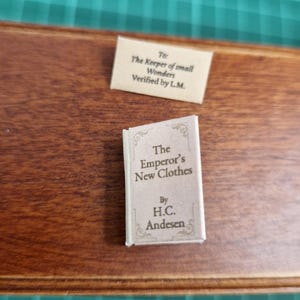 Puede incluir: Un pequeño libro titulado "The Emperor's New Clothes" de H.C. Andersen, con una cubierta color crema y texto. Una pequeña nota arriba dice "To: The Keeper of small Wonders Verified by L.M."