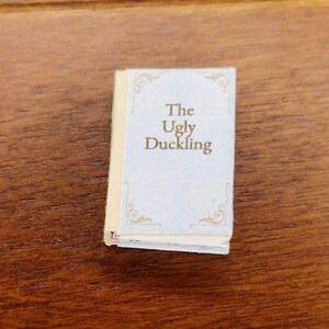 Pode incluir: Um livro em miniatura intitulado "The Ugly Duckling" com uma capa azul clara e letras douradas. O livro tem detalhes dourados decorativos nos cantos e está sobre uma superfície de madeira.