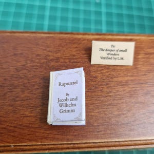 Puede incluir: Un libro en miniatura titulado "Rapunzel" de Jacob y Wilhelm Grimm, con una cubierta clara y texto. Una pequeña nota dice: "To: The Keeper of small Wonders Verified by L.M."