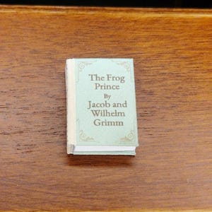 Puede incluir: Un libro en miniatura titulado "The Frog Prince" de Jacob y Wilhelm Grimm. El libro tiene una cubierta verde claro con letras doradas y adornos decorativos. El libro es pequeño, probablemente para una casa de muñecas o exhibición.