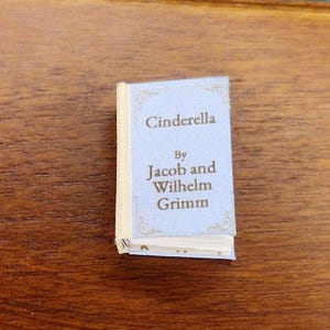 Puede incluir: Un libro en miniatura titulado "Cenicienta" de Jacob y Wilhelm Grimm. El libro tiene una cubierta azul claro con letras doradas y bordes decorativos. El libro está sobre una superficie de madera.