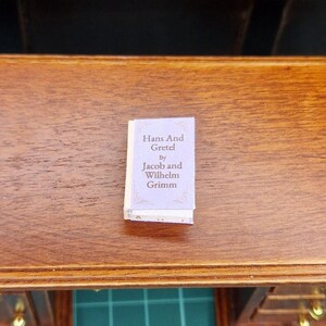 Puede incluir: Un libro en miniatura titulado "Hans And Gretel" de Jacob y Wilhelm Grimm. El libro tiene una cubierta de color lila claro con texto blanco y bordes decorativos. El libro está sobre una superficie de madera.