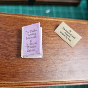 Puede incluir: Un libro en miniatura titulado "The Twelve Dancing Princesses" de Jacob y Wilhelm Grimm, con una cubierta lavanda y texto blanco. Junto a él hay una pequeña tarjeta que dice "The Keeper of small Wonders, Verified by L.M."
