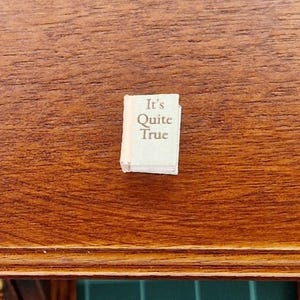 Puede incluir: Un libro en miniatura con el título "It's Quite True" en una fuente serif. El libro es blanco con una cubierta de color claro y está sobre una superficie de madera. La madera tiene un tono marrón cálido.