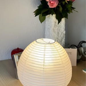 Könnte beinhalten: Eine runde, weiße Papierlaterne mit Metallfuß steht auf einer Holzoberfläche. Im Hintergrund eine Vase mit rosa Rosen. Ein roter Apfel und ein gerahmtes Bild sind ebenfalls zu sehen.