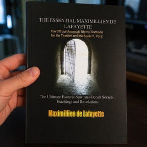 Op de afbeelding: Een zwart boek met de titel The Essential Maximillien de Lafayette, The Official Anunnaki Ulema Textbook for the Teacher and the Student. Vol.2. De omslag bevat een afbeelding van een open poort die naar fel licht leidt en witte tekst.
