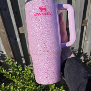 Peut inclure: Un gobelet Stanley rose paillet&eacute; avec une poign&eacute;e. Le gobelet a un couvercle et une poign&eacute;e rose clair, et le logo Stanley est en rose. Le gobelet est tenu devant une cl&ocirc;ture en bois et un feuillage vert.