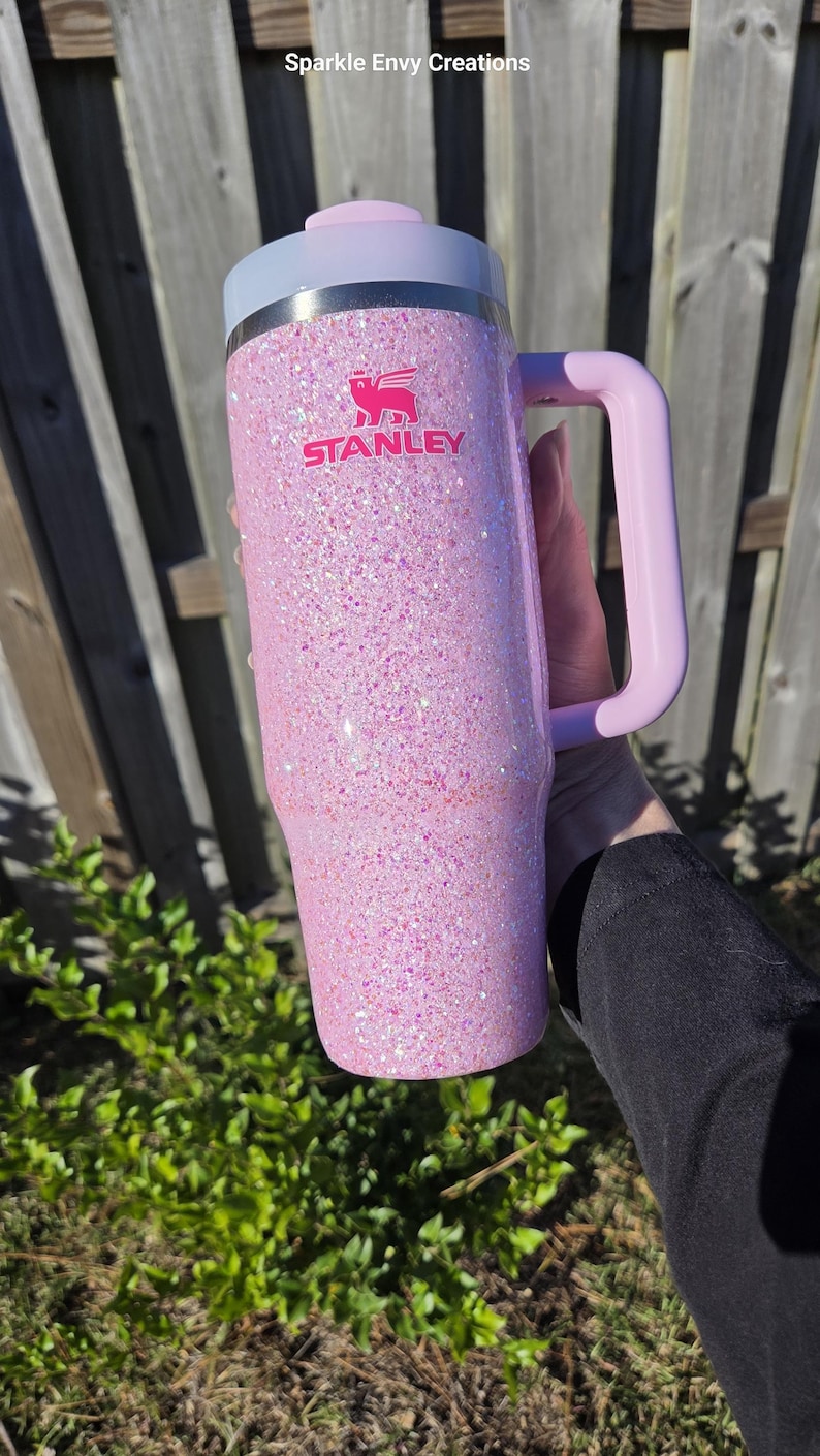 Peut inclure: Un gobelet Stanley rose paillet&eacute; avec une poign&eacute;e. Le gobelet a un couvercle et une poign&eacute;e rose clair, et le logo Stanley est en rose. Le gobelet est tenu devant une cl&ocirc;ture en bois et un feuillage vert.