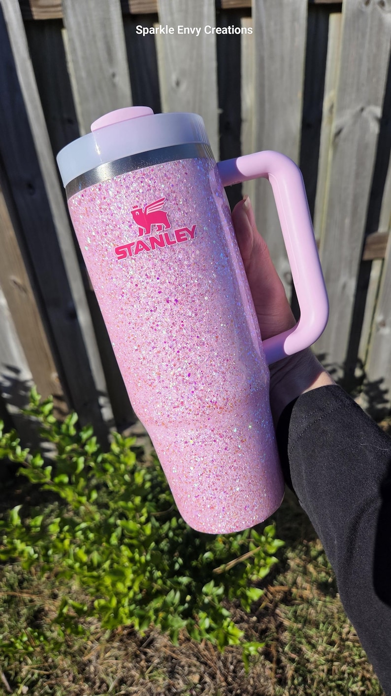 Peut inclure: Un gobelet Stanley rose paillet&eacute; avec une poign&eacute;e et un couvercle rose clair. Le logo Stanley est en rose. Le gobelet est tenu par une personne. Le texte "Sparkle Envy Creations" est en arri&egrave;re-plan.
