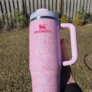 Peut inclure: Un mug Stanley rose paillet&eacute; avec une poign&eacute;e et un couvercle rose clair. Le mug porte le logo Stanley en rose. L'arri&egrave;re-plan est une sc&egrave;ne ext&eacute;rieure floue avec une cl&ocirc;ture en bois et de l'herbe verte.