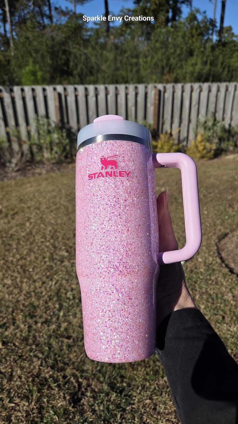 Peut inclure: Un mug Stanley rose paillet&eacute; avec une poign&eacute;e et un couvercle rose clair. Le mug porte le logo Stanley en rose. L'arri&egrave;re-plan est une sc&egrave;ne ext&eacute;rieure floue avec une cl&ocirc;ture en bois et de l'herbe verte.