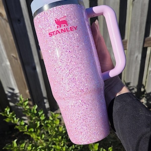 Peut inclure: Un gobelet Stanley rose paillet&eacute; avec une poign&eacute;e. Le gobelet a un couvercle et une poign&eacute;e rose clair, avec le logo Stanley en rose. Le gobelet est tenu devant une cl&ocirc;ture en bois et un feuillage vert.