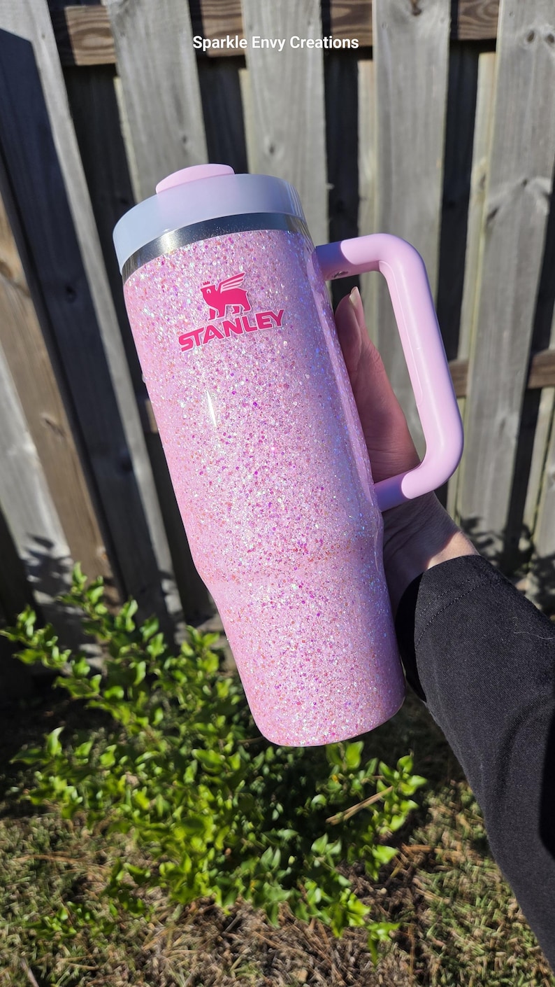 Peut inclure: Un gobelet Stanley rose paillet&eacute; avec une poign&eacute;e. Le gobelet a un couvercle et une poign&eacute;e rose clair, avec le logo Stanley en rose. Le gobelet est tenu devant une cl&ocirc;ture en bois et un feuillage vert.