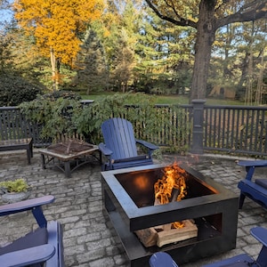 May include: Outdoor patio scene with a square metal fire pit containing a burning fire. Four blue Adirondack chairs surround the fire pit on a brick patio. Trees with yellow and green foliage are in the background.