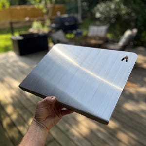 May include: A rectangular, brushed metal grilling surface with a handle cutout. The metal has a reflective quality, and the edges are dark. The item is held up in front of a blurred outdoor background.