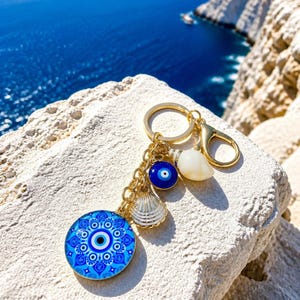 Könnte beinhalten: Ein goldfarbener Schlüsselanhänger mit einem runden blau-weißen Nazar-Amulett mit Mandalamuster, einer kleinen blauen Nazar-Perle, einem weißen Muschelanhänger und einer glatten weißen Muschel, befestigt an einem goldenen Metallring und Karabiner.
